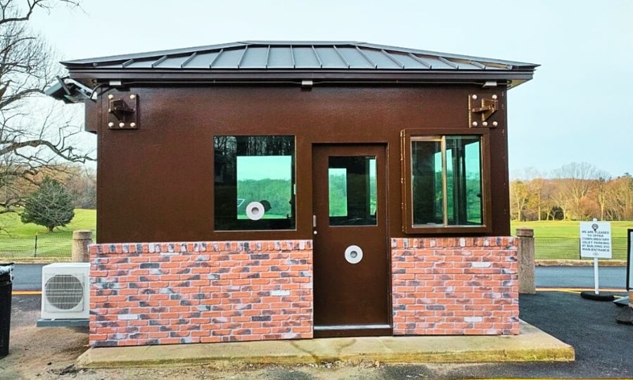 Security guard booth with a brick base and steel upper structure, featuring ballistic glass windows and a central bulletproof door, situated outdoors for access control.